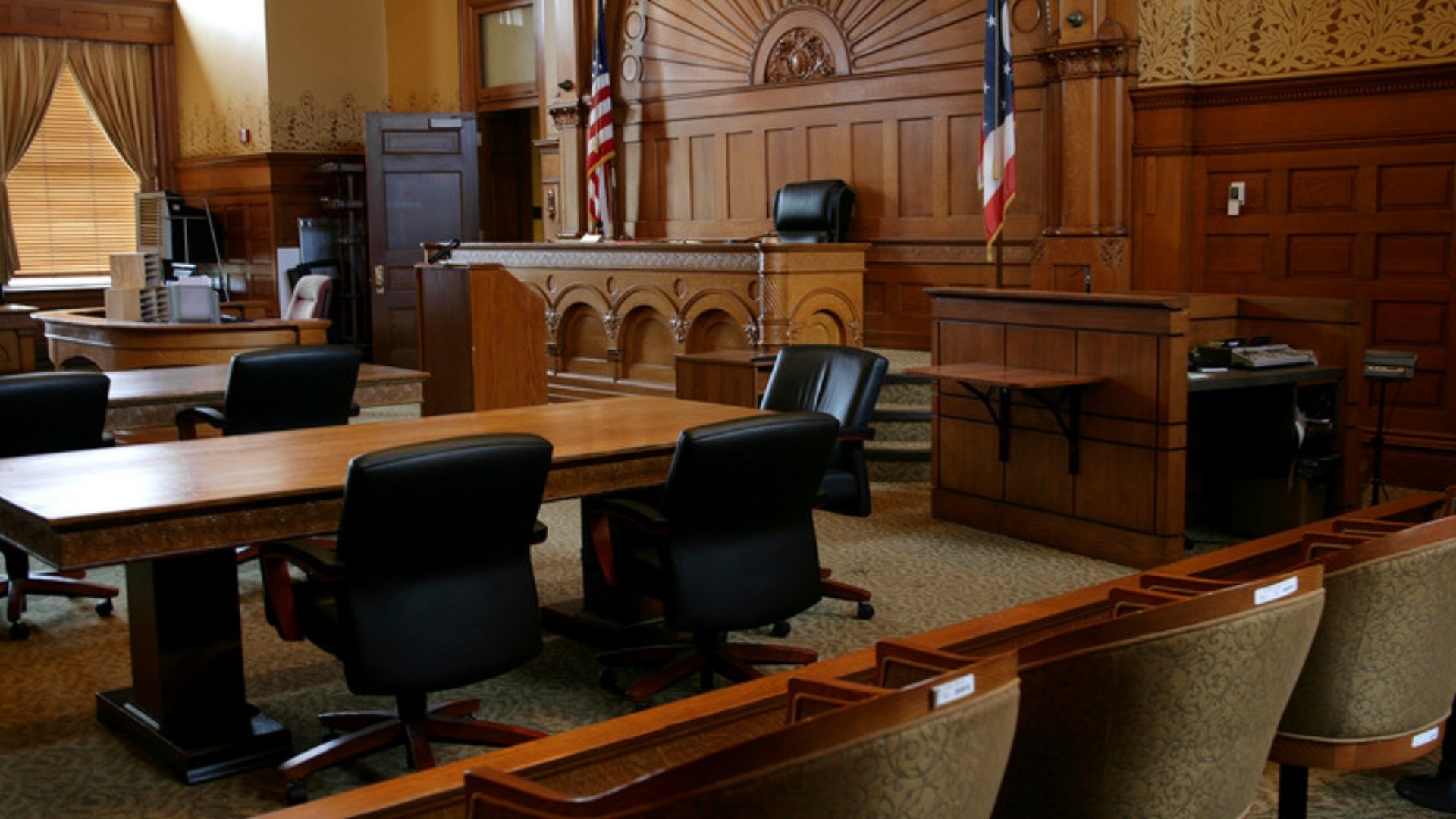 An empty wood-paneled courtroom with counsel tables, a judge's bench and two flags.
