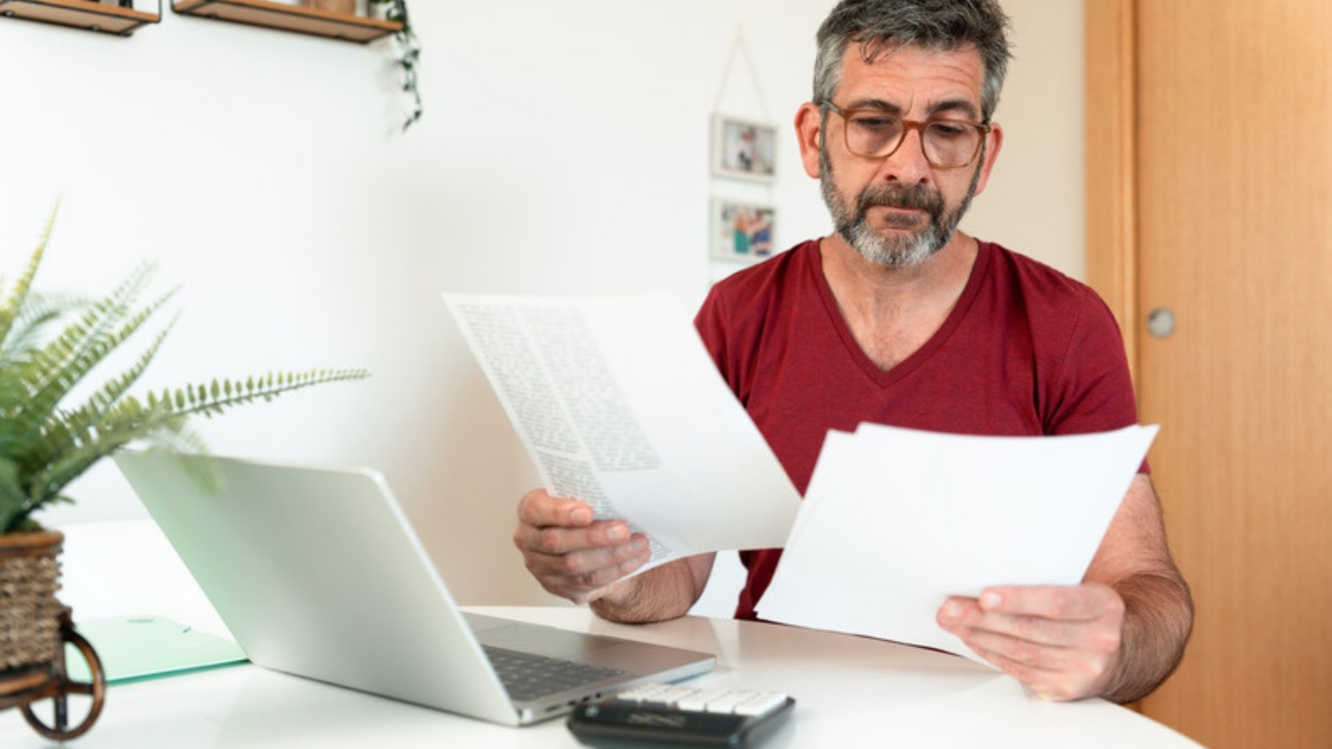 A man in glasses sits at a white desk reading two printed pages beside a laptop.
